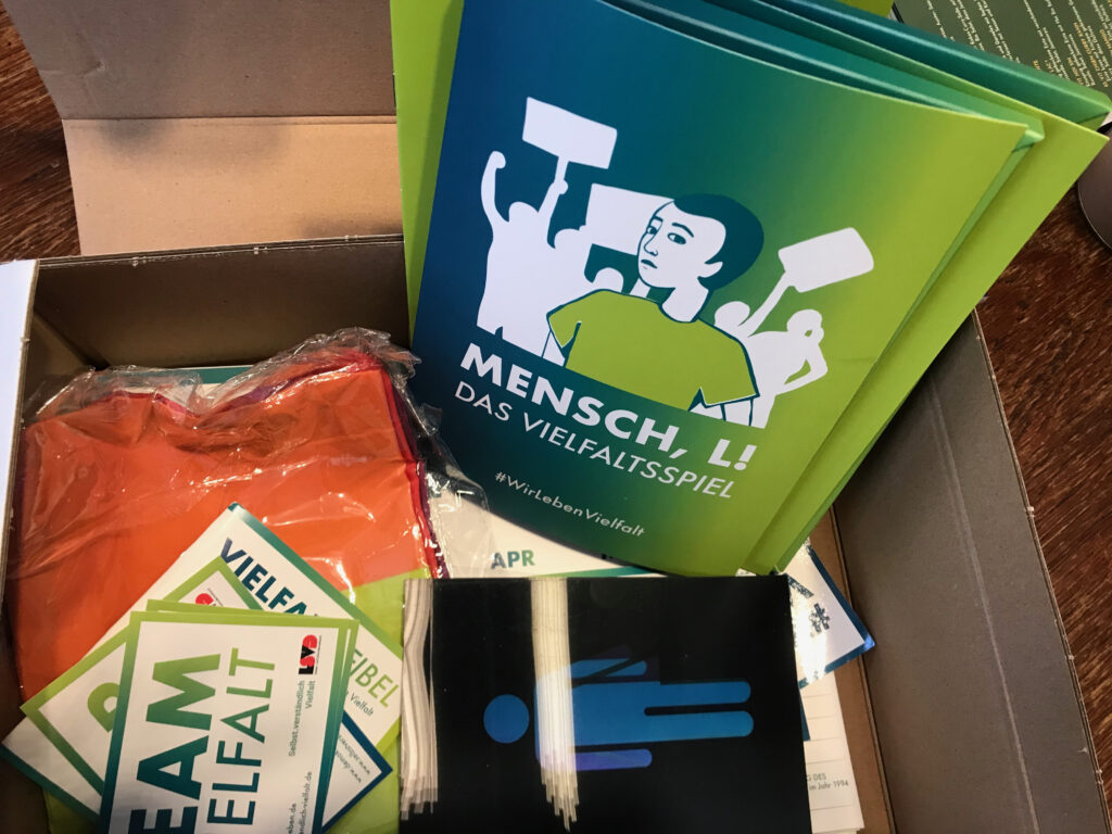 Zu sehen ist ein Karton mit der Aufschrift "Queer-Paket für die Jugendarbeit" in grün/gelb und auf dem zweiten Bild der Inhalt des Kartons, Flyer und Broschüre, eine Fahne und Aufkleber.